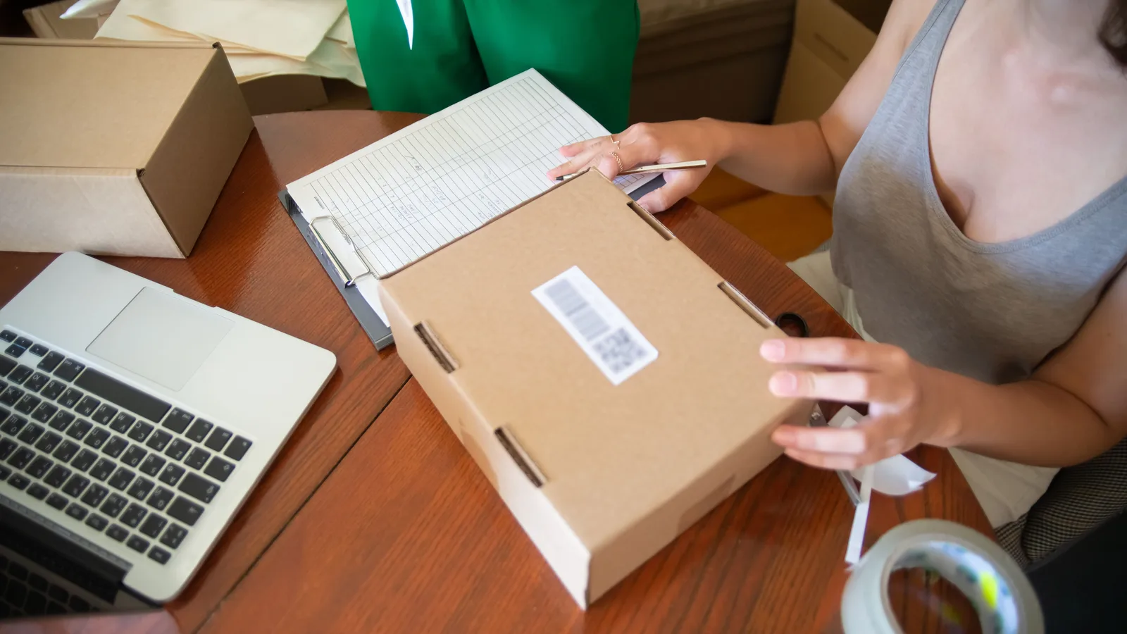Person checking paperwork while preparing a parcel beside a laptop