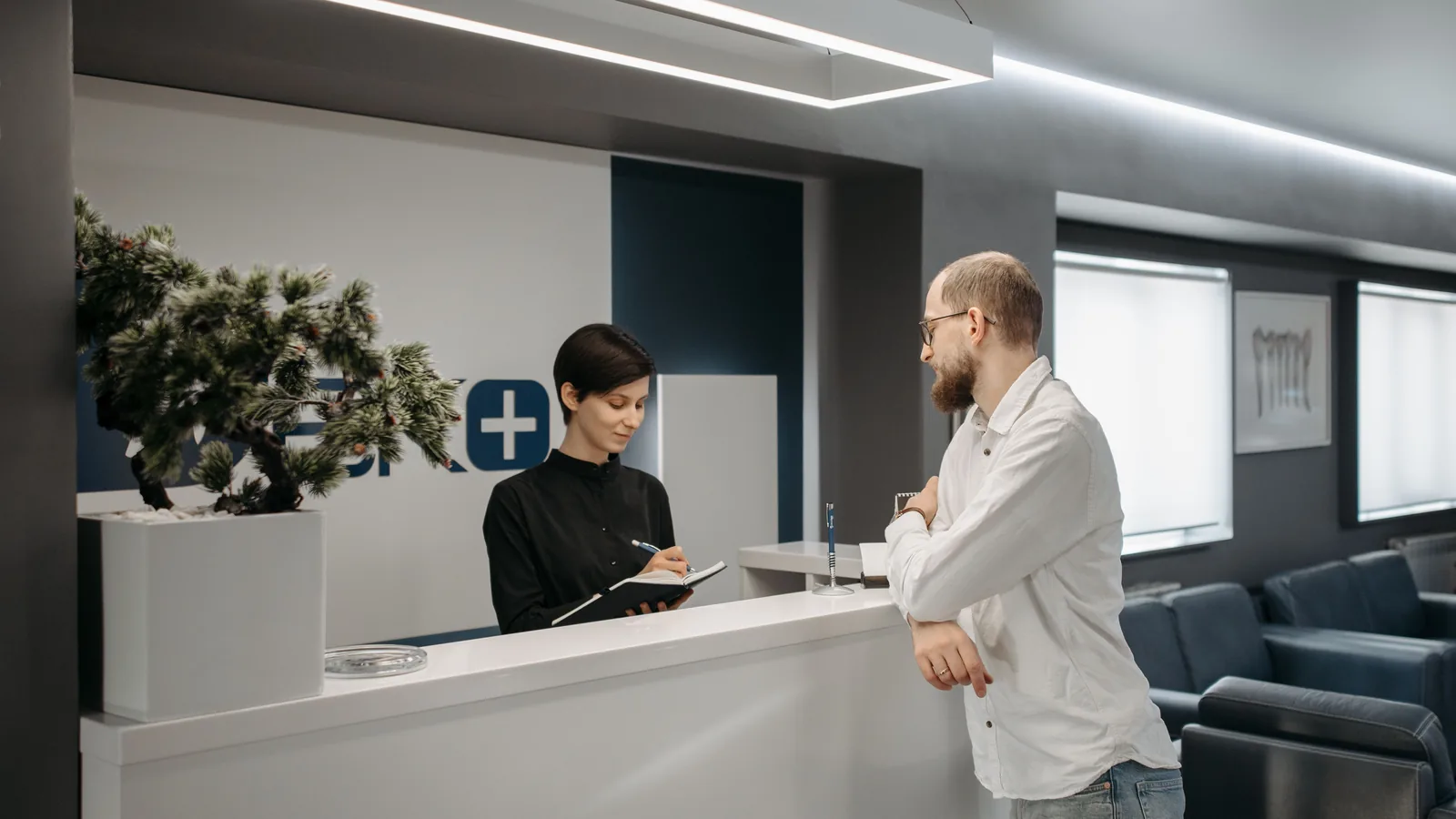 Receptionist writing in a notebook while speaking with a visitor at a clinic desk