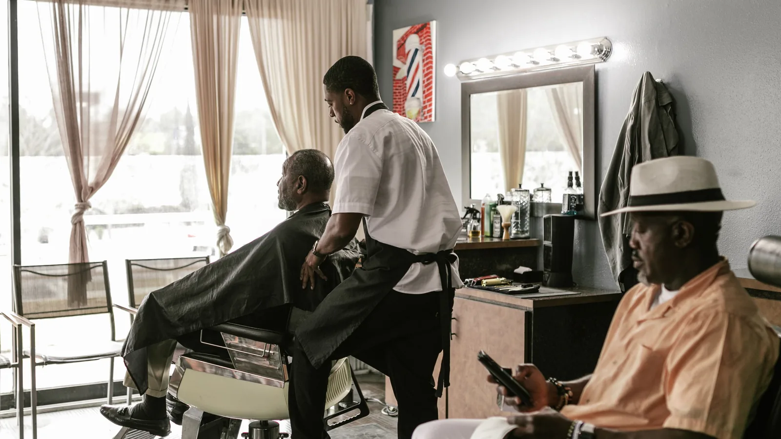 Barber beside a customer in a barber chair, with another seated customer nearby