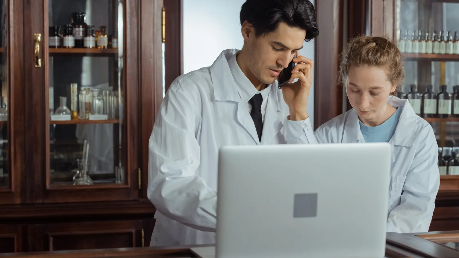 Two pharmacists working together at a laptop beside stocked shelves